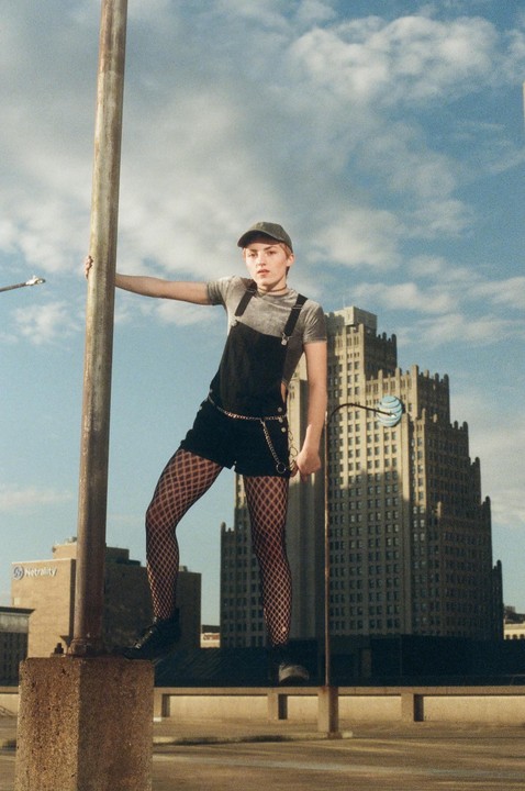 A woman in a baseball cap and overalls poses on a concrete pillar with a city skyline in the background.