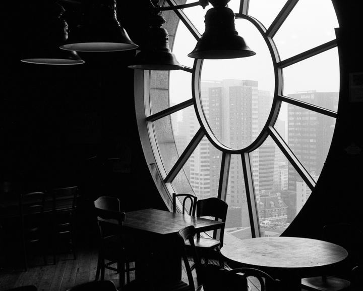 A dark room with tables and chairs is illuminated by a large, round window looking out onto a city.