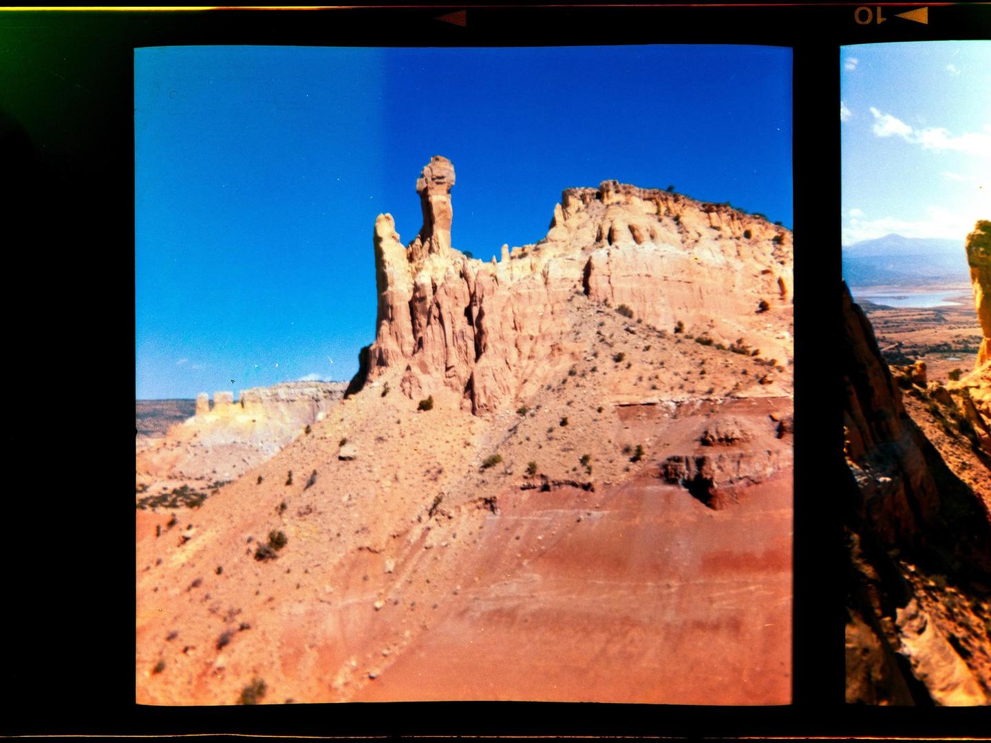 A tall, narrow rock formation stands prominently against a clear blue sky in a desert landscape.