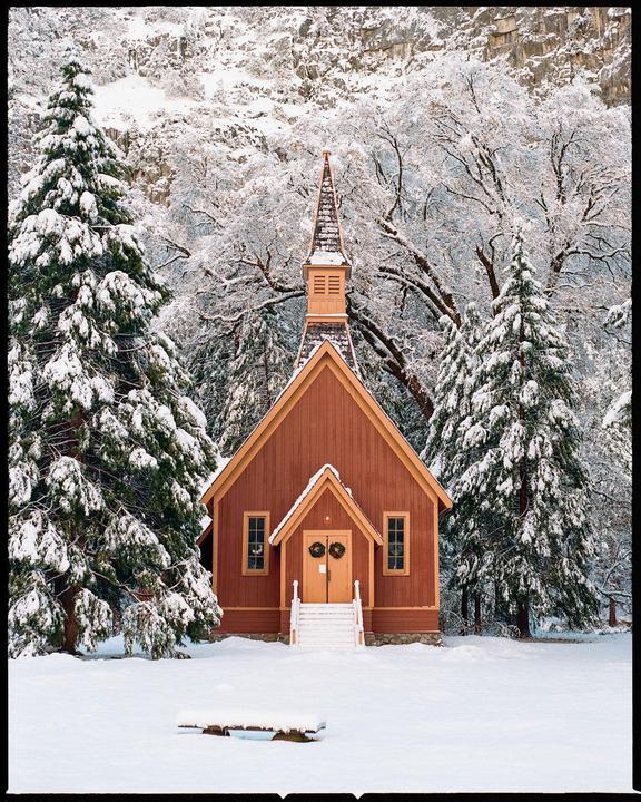A small red chapel with a steeple stands in a snowy landscape surrounded by snow-covered trees.