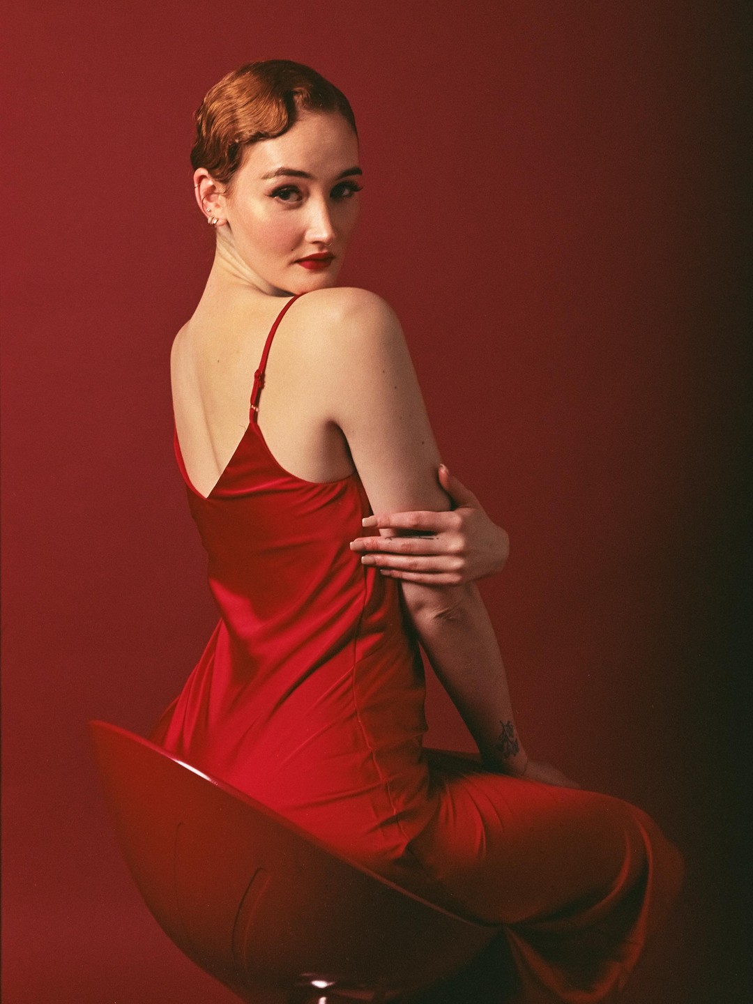 A woman in a red dress with a vintage hairstyle sits on a red chair against a red background.