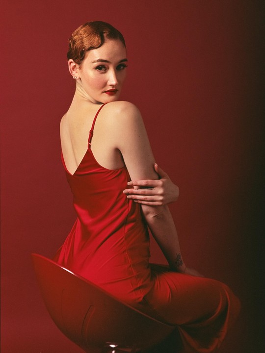 A woman in a red dress with a vintage hairstyle sits on a red chair against a red background.