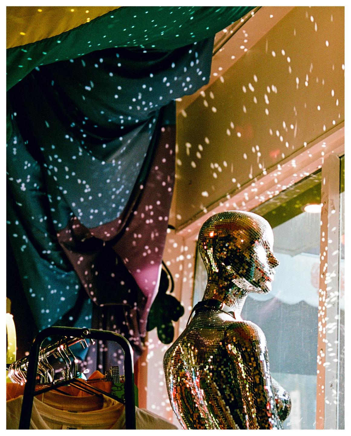A disco ball mannequin stands in a window display, reflecting light onto the surrounding walls and fabric.
