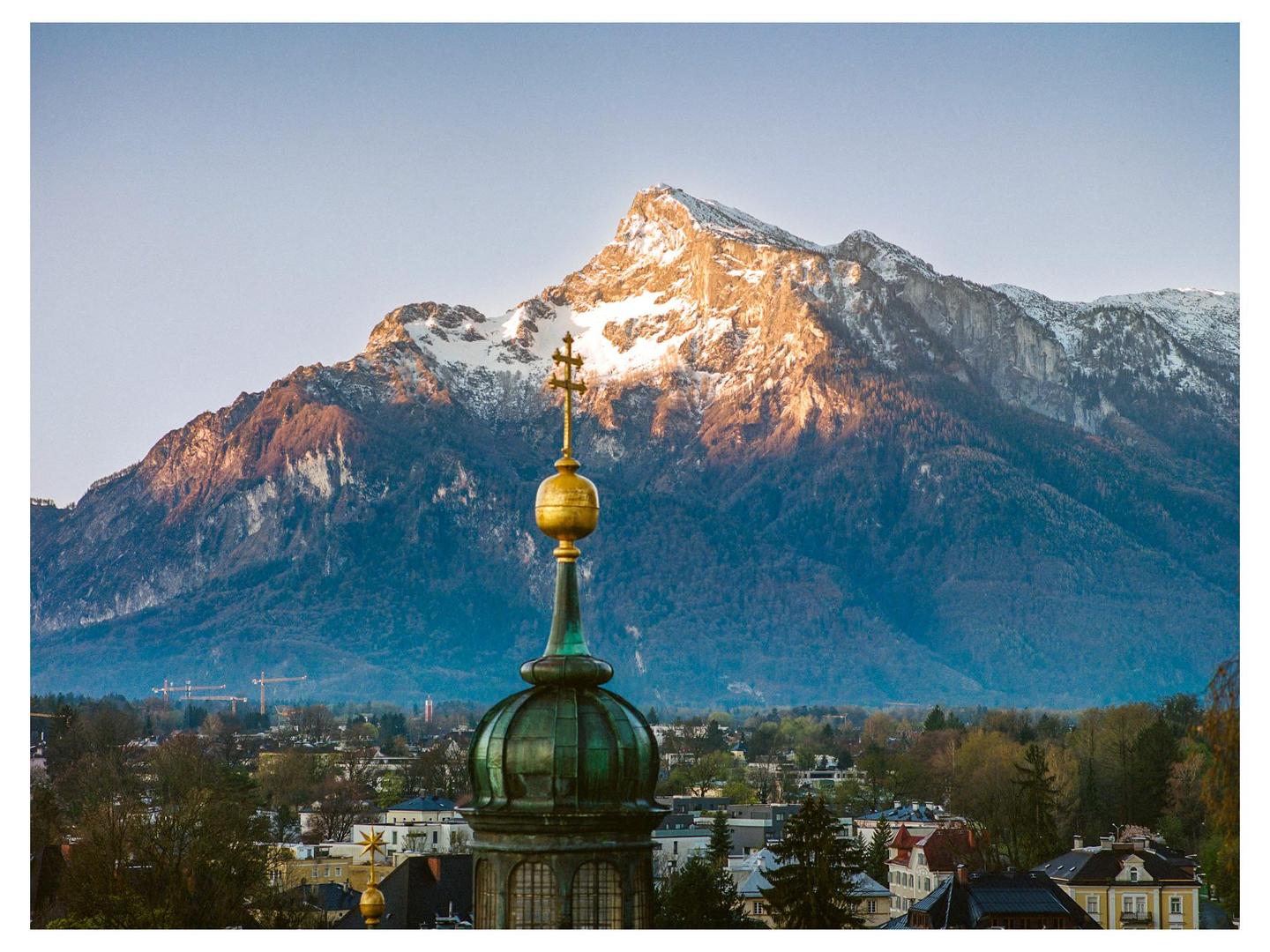 A golden church spire rises above a town with snow-capped mountains in the background.