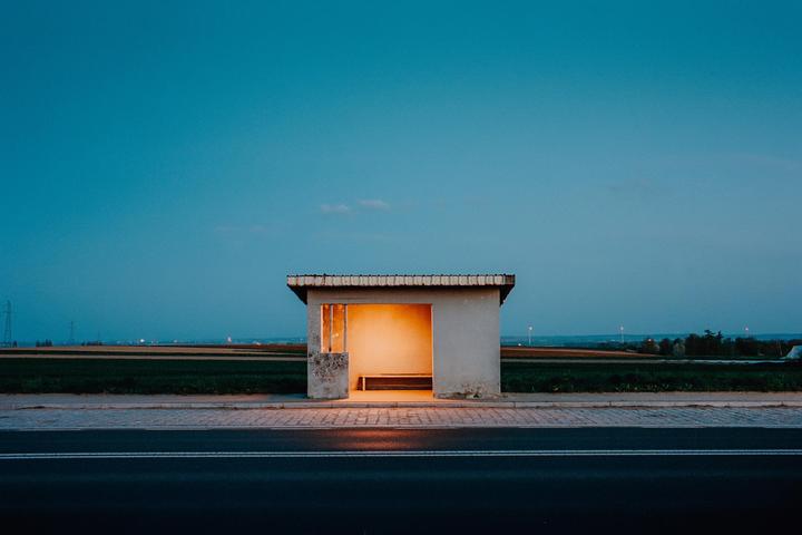 A bus stop with an illuminated interior sits beside a road at dusk, with fields and a blue sky in the background.