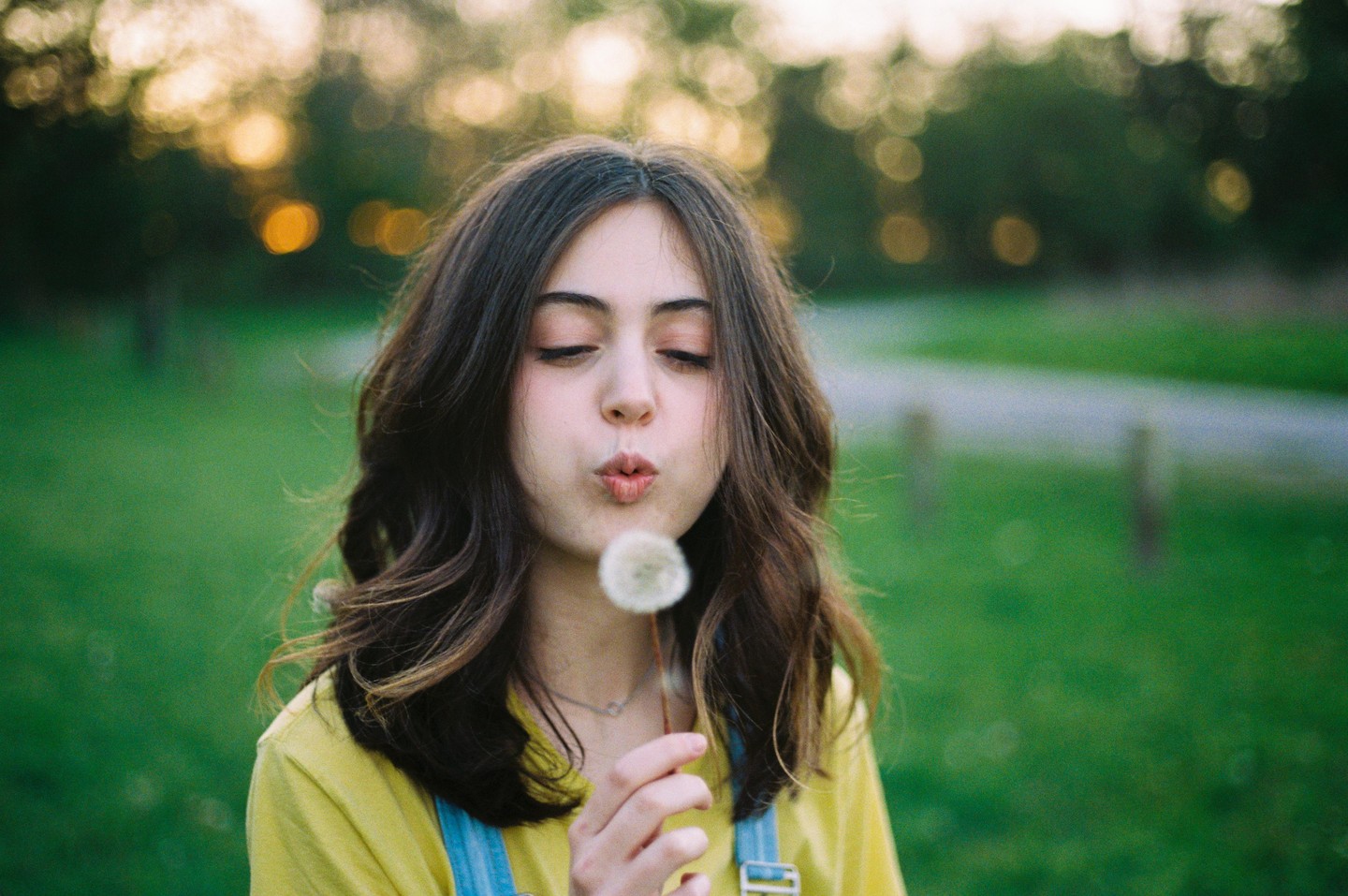 A young woman with her eyes closed blows on a dandelion in a grassy field.