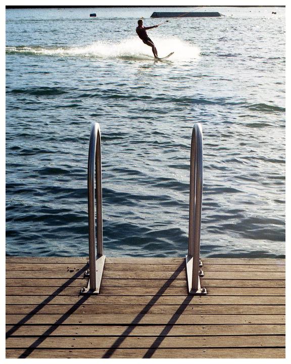 A person water skis on a lake with a wooden dock and metal railings in the foreground.