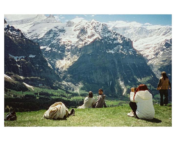 A group of people are sitting on a grassy hill overlooking a valley with snow-capped mountains.