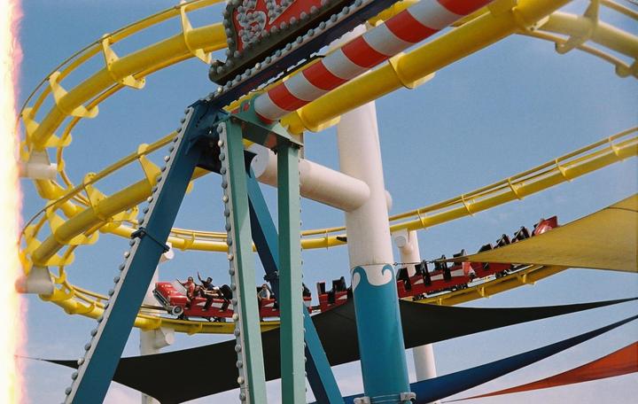 A red roller coaster car with people riding in it moves along a yellow track against a clear blue sky.