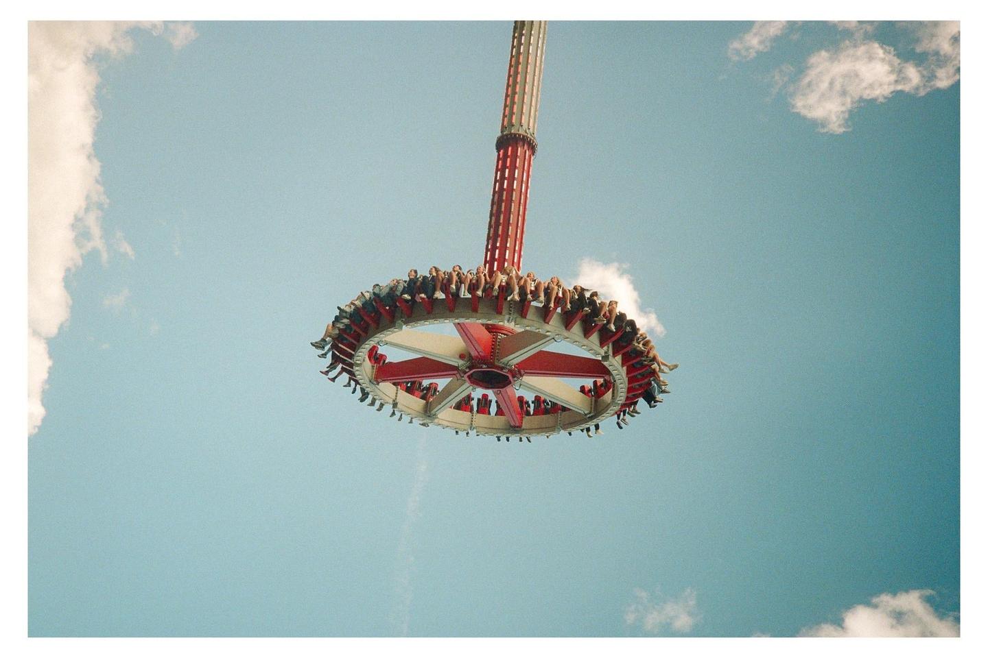 A large amusement park ride with people seated around its circular edge is suspended high in the air against a blue sky with white clouds.