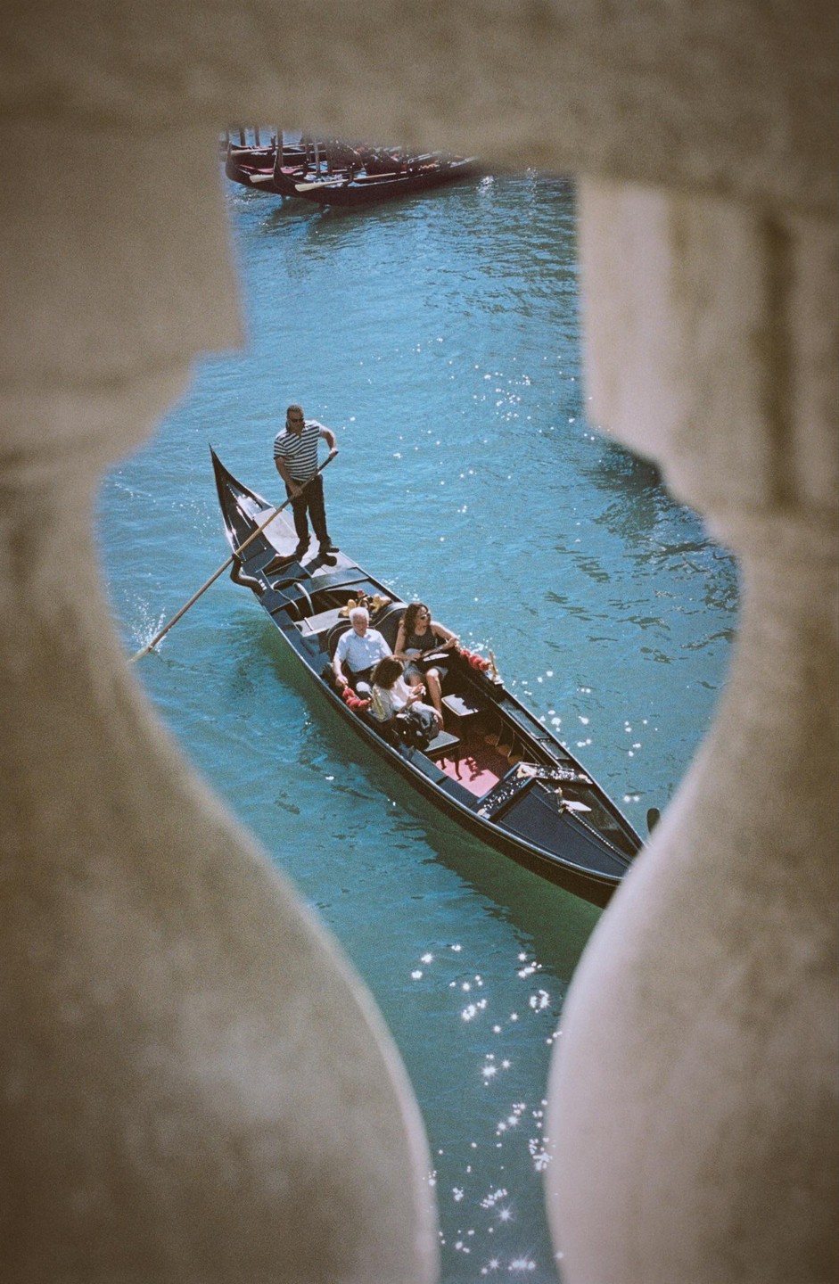A gondola with a gondolier and three passengers is seen from above through an archway on a sunny day in Venice.
