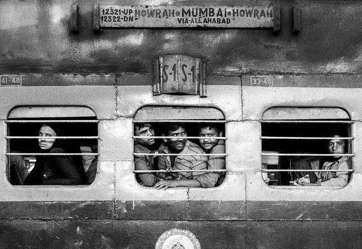 Several people look out from the barred windows of a train car with a destination sign for Mumbai and Howrah.