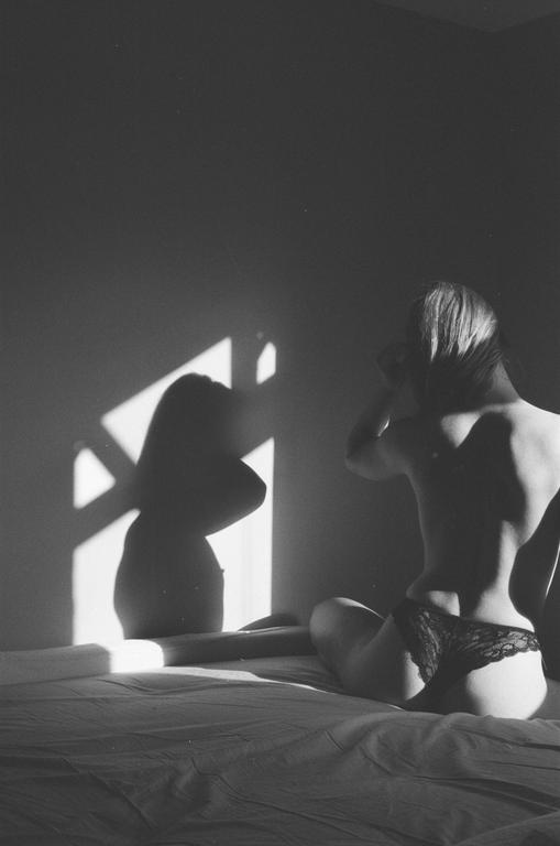 A woman sits on a bed with her back to the viewer, looking at her shadow on the wall.