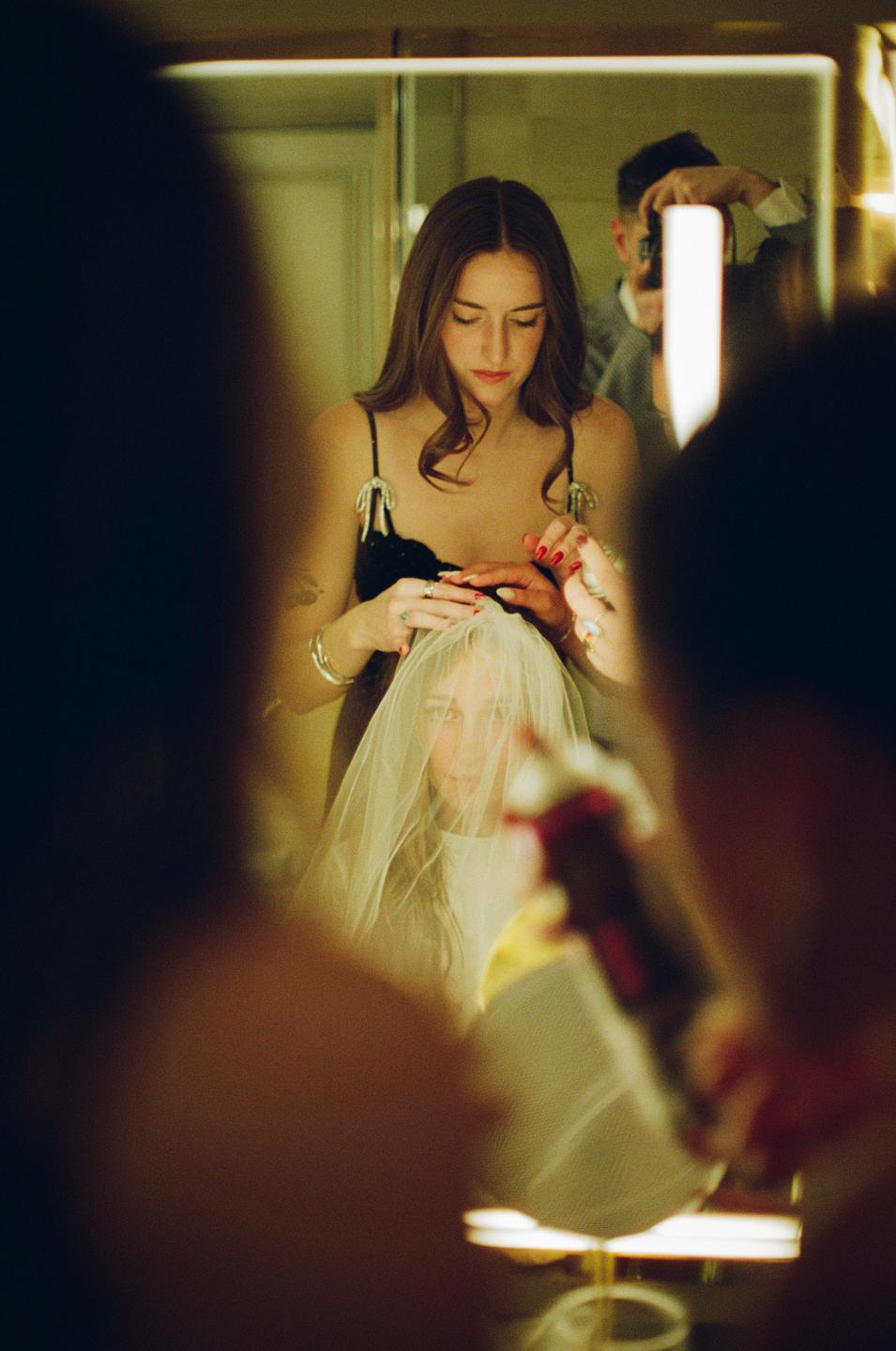 A woman in a black dress adjusts a veil on another woman in a mirror reflection.