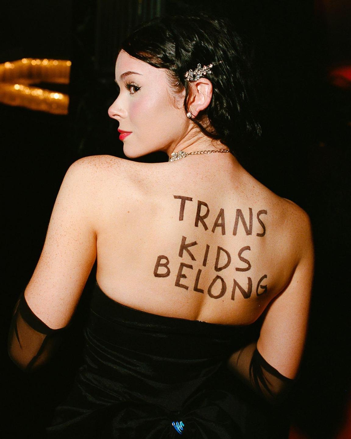 A woman with dark hair and red lipstick looks over her shoulder, revealing the phrase "TRANS KIDS BELONG" written on her back.