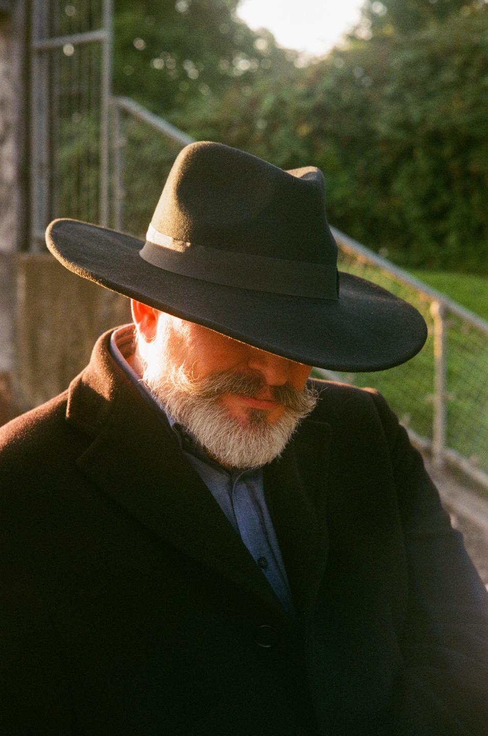 A man with a beard and mustache wears a hat and a dark coat, with sunlight illuminating his face.