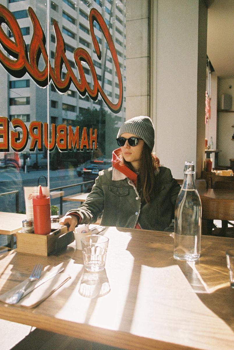 A woman wearing a beanie and sunglasses sits at a diner table with a bottle of water and condiments, with a city street visible through the window.