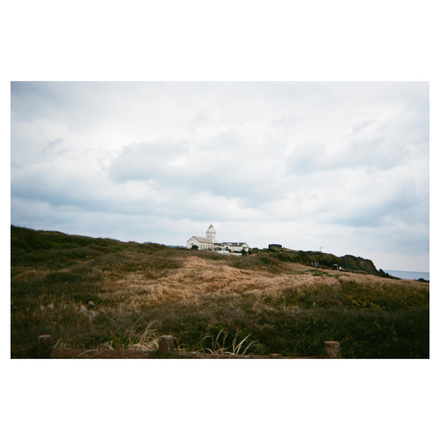 A white building with a tower sits on a grassy hill under a cloudy sky.
