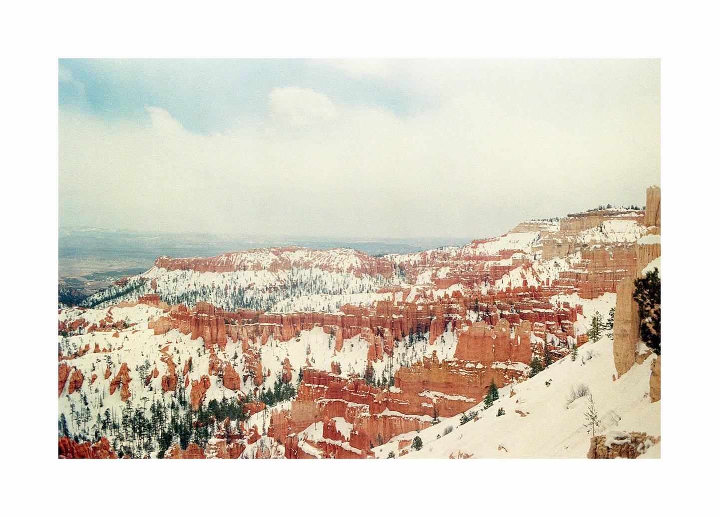 A vast canyon landscape with red rock formations is covered in snow under a cloudy sky.