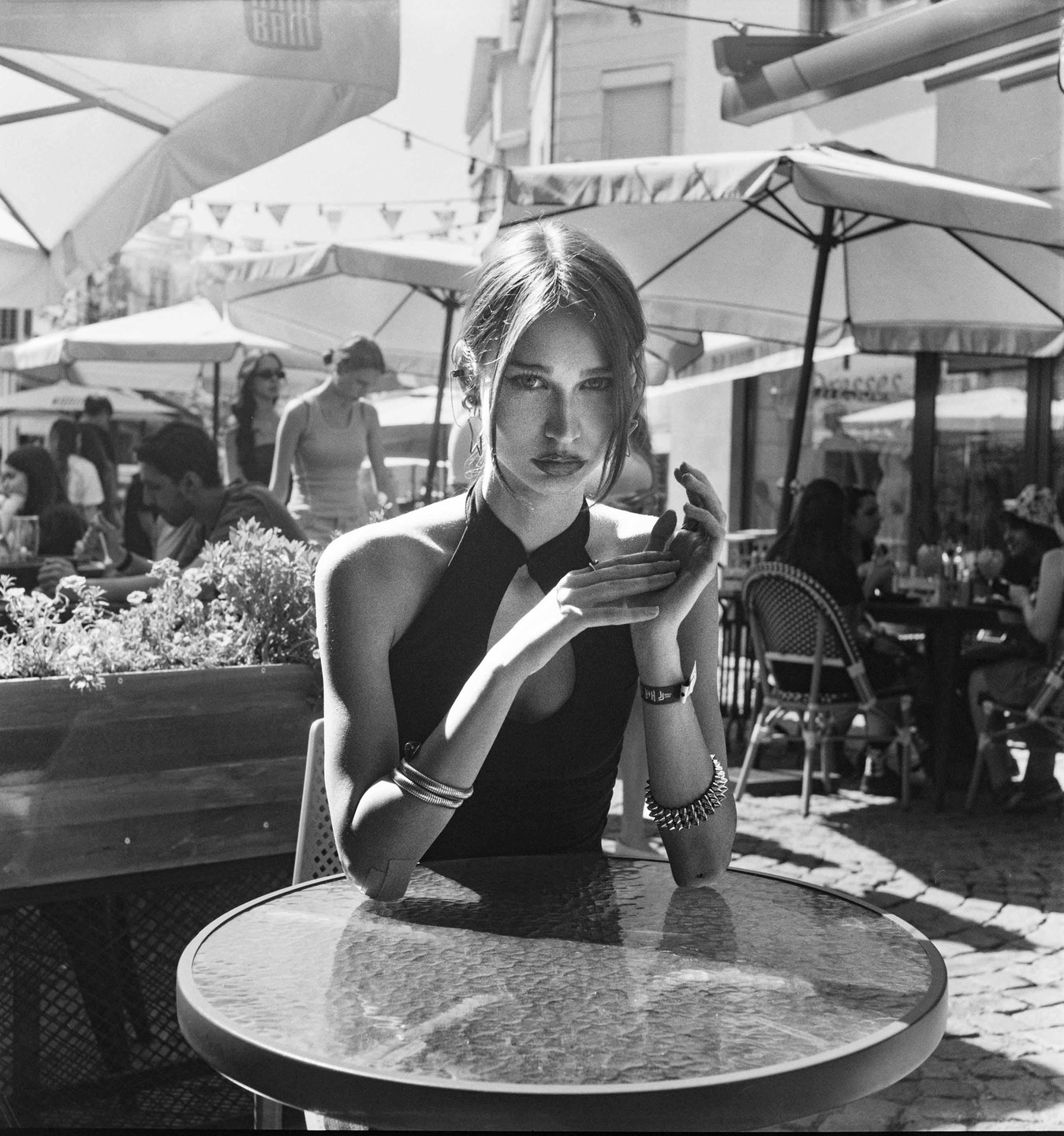 A woman in a dark dress sits at a table in an outdoor cafe with other patrons and umbrellas in the background.