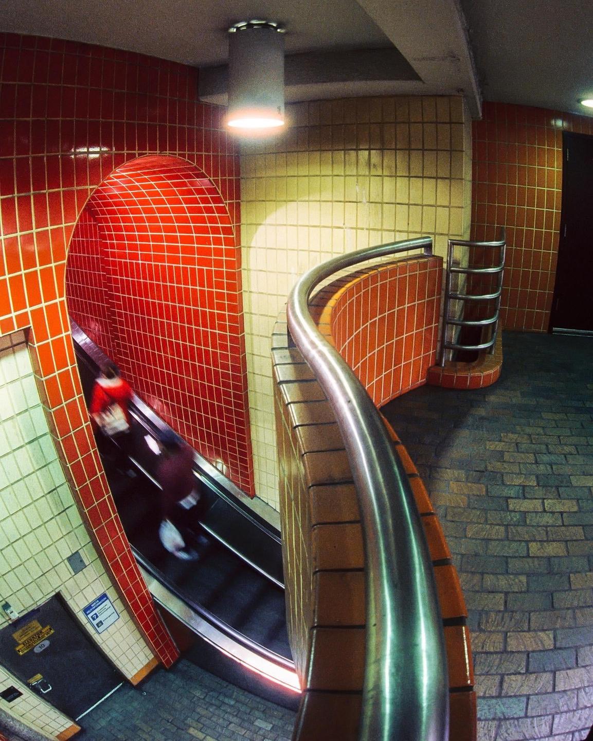 A person rides an escalator down into a subway station with red and yellow tiled walls.