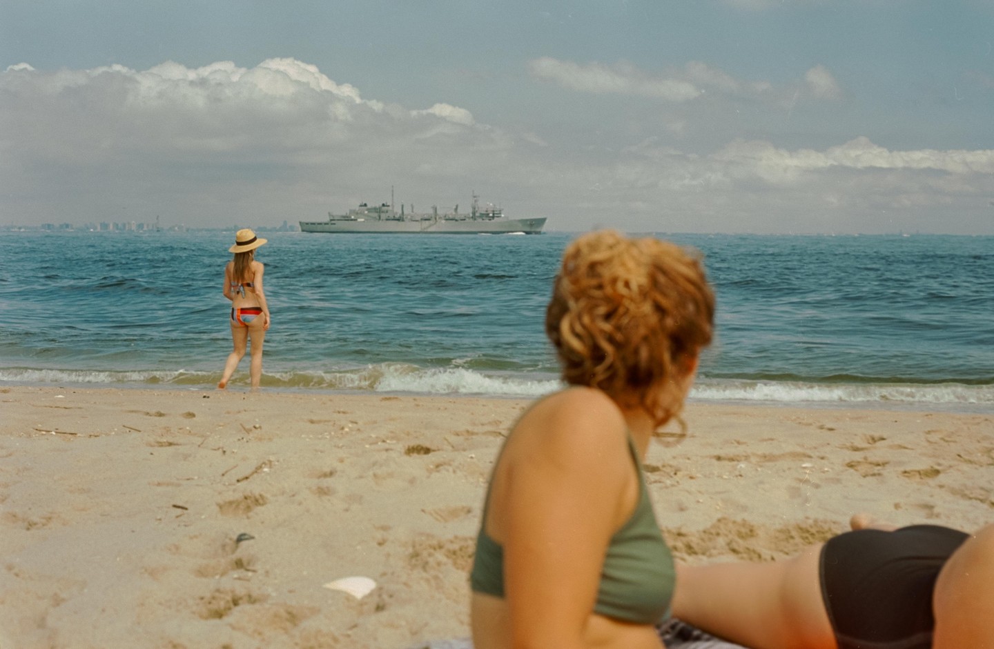 A woman in a straw hat and bikini stands in the ocean looking at a ship while another woman sits on the sand in the foreground.