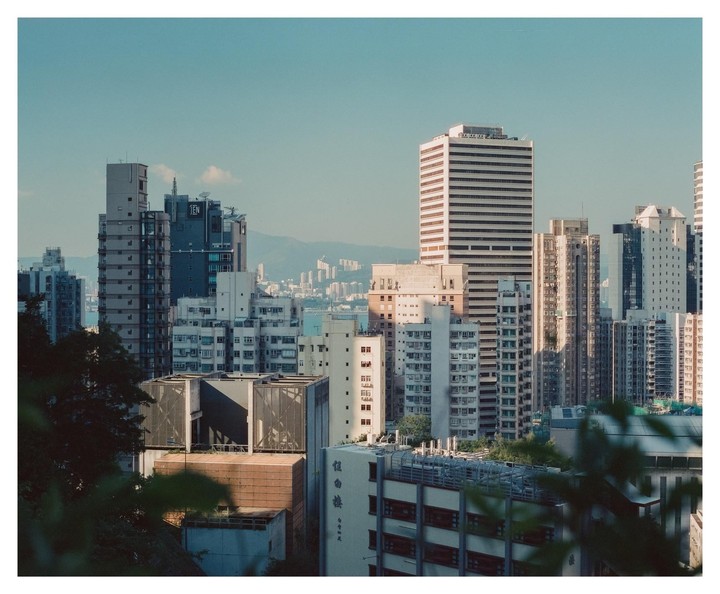 A dense urban landscape of Hong Kong features numerous tall buildings under a clear sky, with mountains visible in the distance.