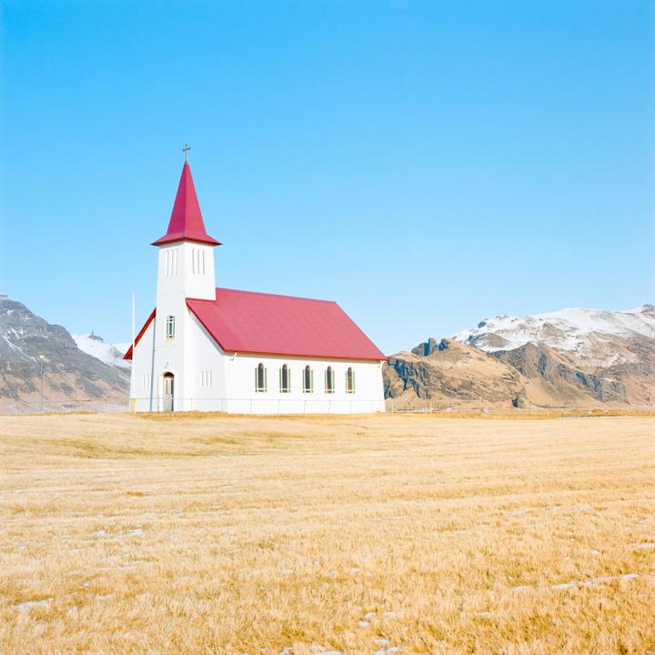 A white church with a red roof stands in a field with mountains in the background.