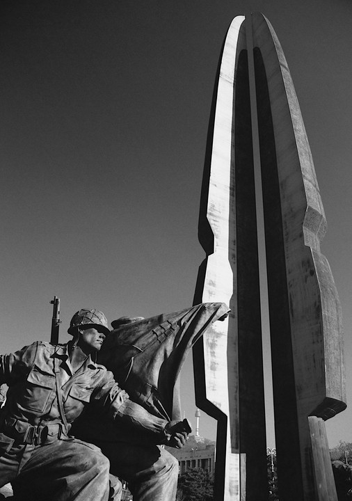 A statue of a soldier holding a rifle and a flag is positioned in front of a tall, abstract monument.