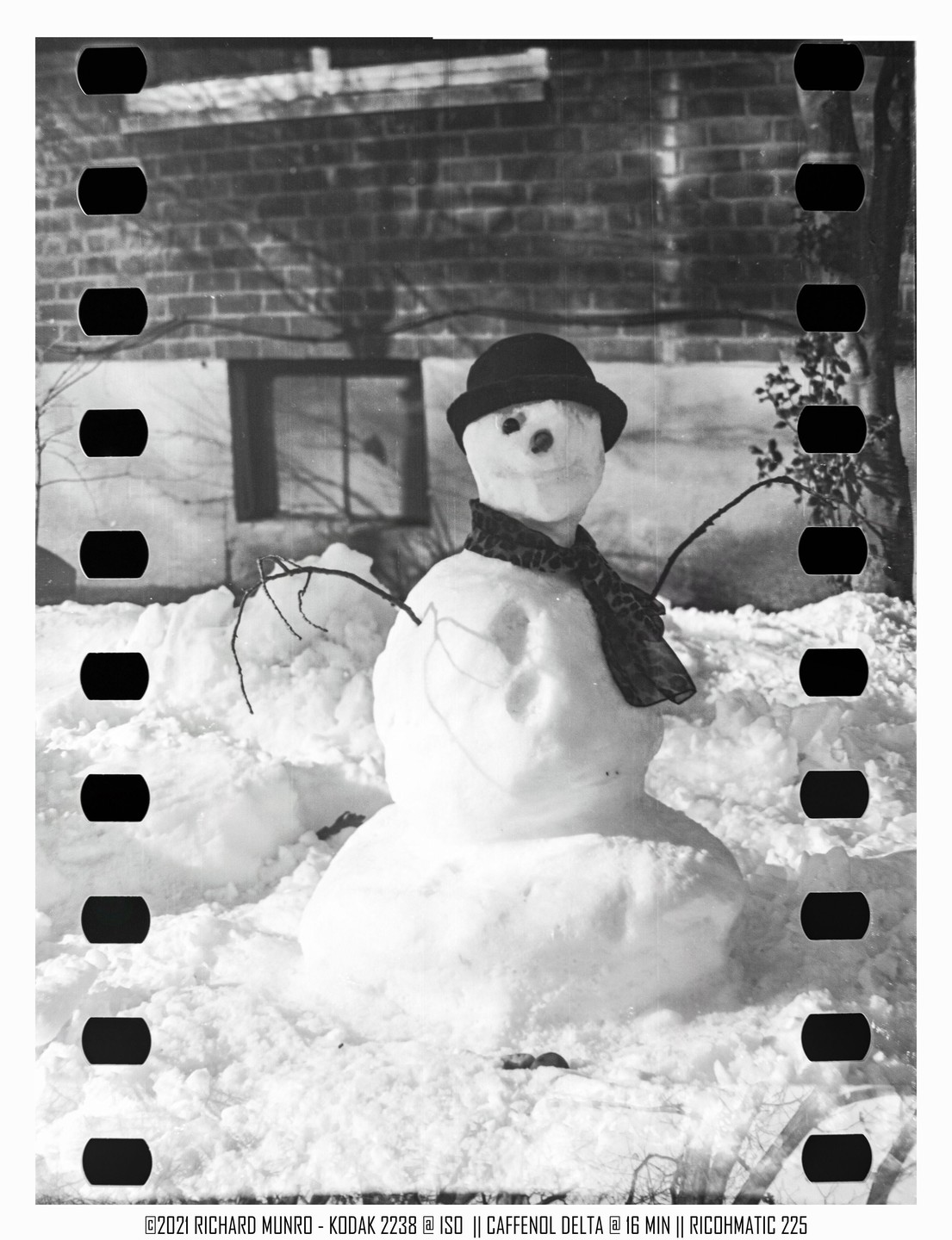 A snowman wearing a hat and scarf stands in a snowy yard in front of a brick building.
