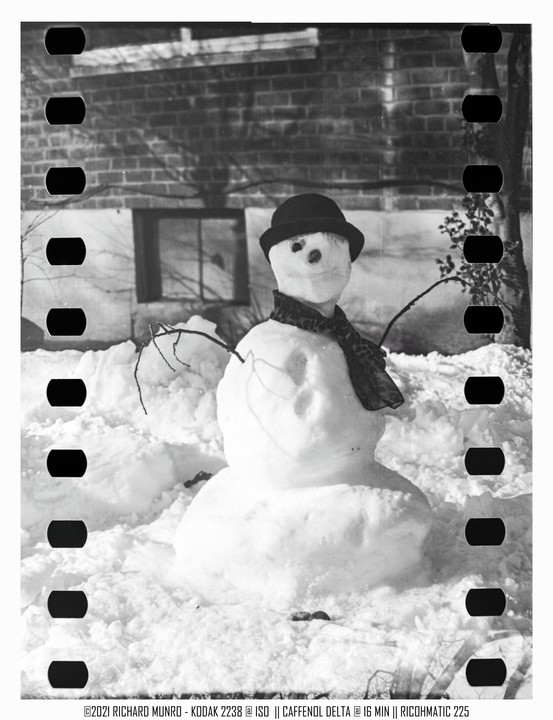 A snowman wearing a hat and scarf stands in a snowy yard in front of a brick building.