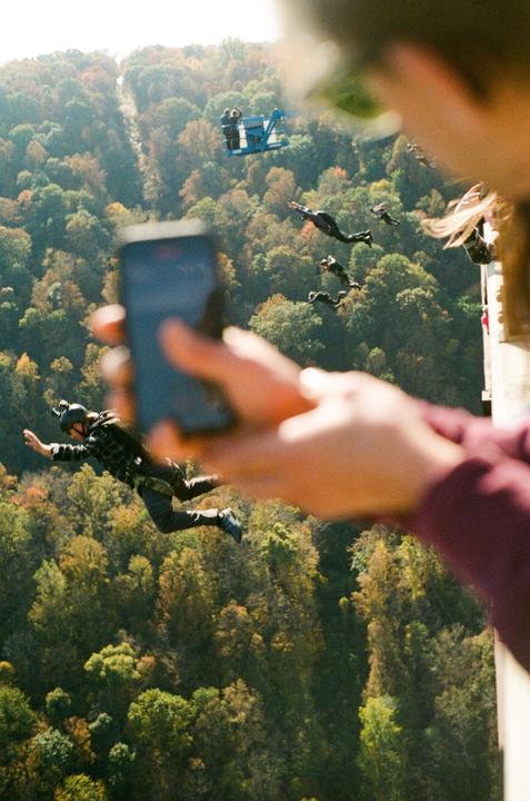 A person holds a phone to record other people falling through the air above a forest.
