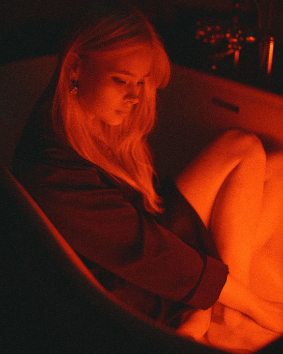 A woman with long hair sits in a bathtub, illuminated by red light.
