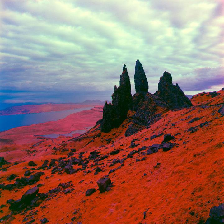 A landscape shows a rocky hillside with prominent rock formations under a cloudy sky, with a body of water and distant land visible.