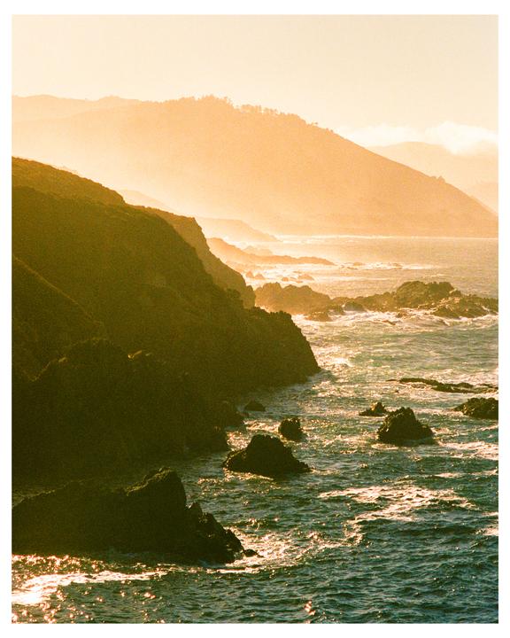 The sun shines on a rocky coastline with mountains in the background.