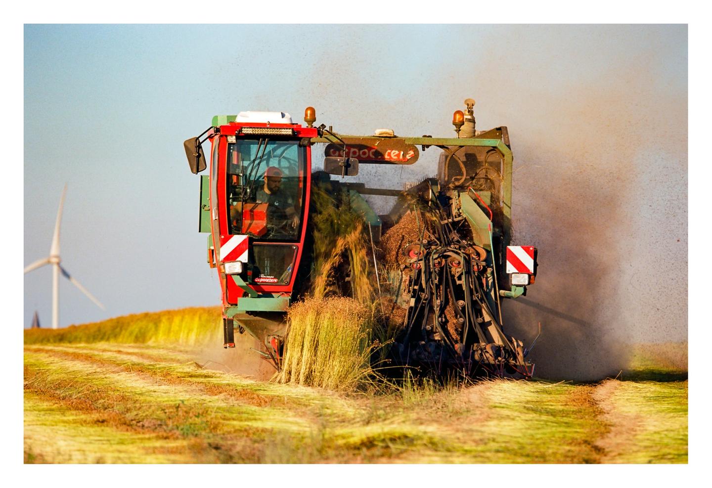 A man operates a combine harvester in a field with a wind turbine in the background.
