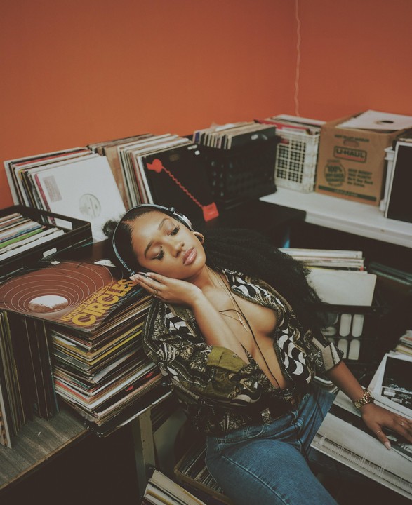 A woman with headphones rests her head on a stack of vinyl records.