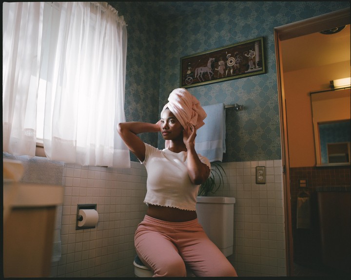 A woman with a towel wrapped around her head sits in a bathroom.
