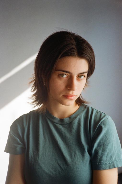 A woman with short hair and a green shirt looks directly at the viewer, with a diagonal shadow on the wall behind her.