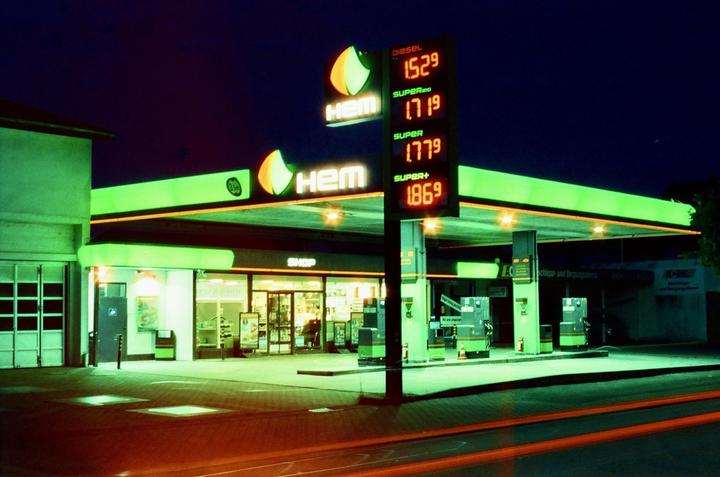 A gas station with bright green and orange lights illuminates the night, with a price sign displaying fuel costs.