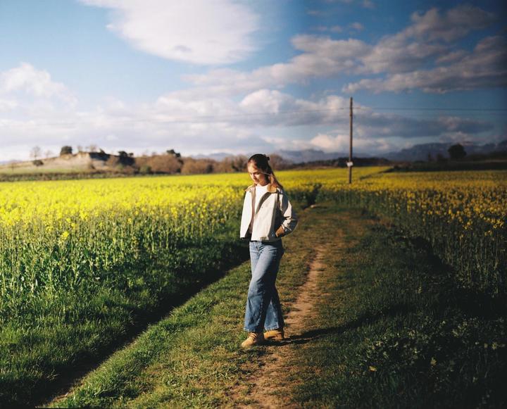 A woman stands on a dirt path through a field of yellow flowers under a partly cloudy sky.