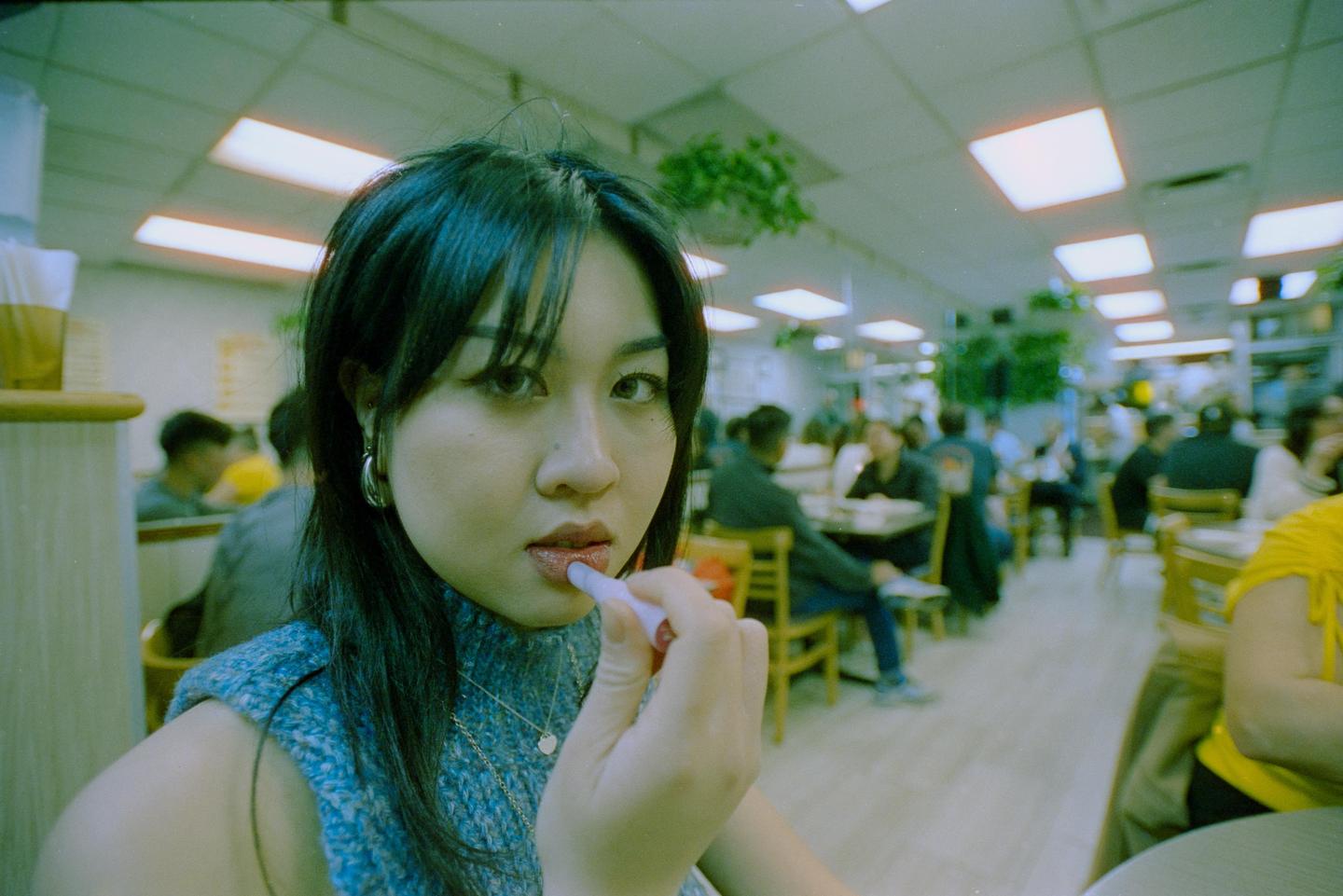 A woman applies lip gloss while looking at the camera in a busy restaurant.
