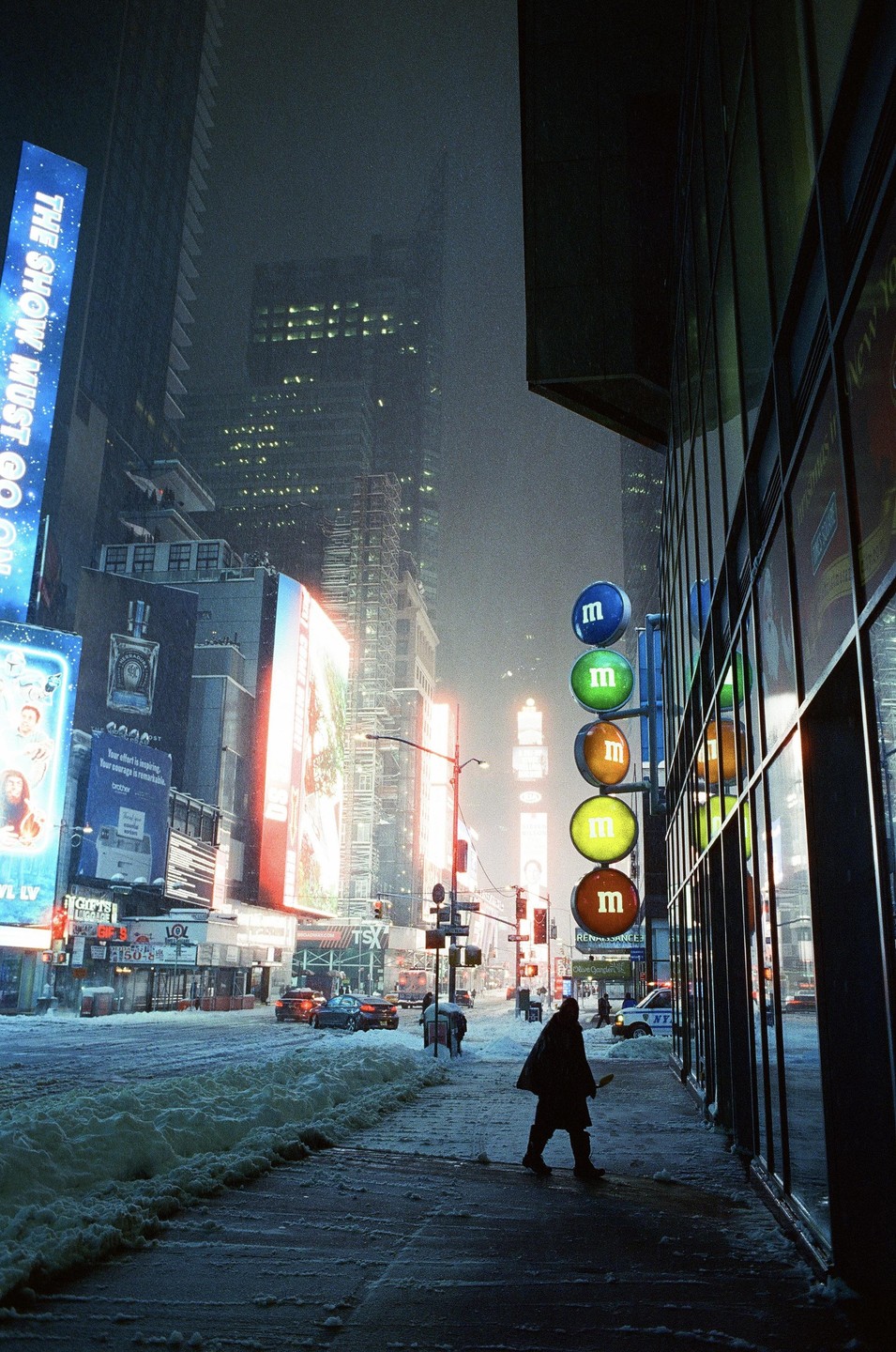 A person walks on a snowy sidewalk next to a building with large round M&M signs in Times Square at night.