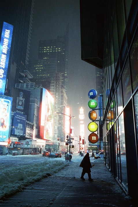 A person walks on a snowy sidewalk next to a building with large round M&M signs in Times Square at night.
