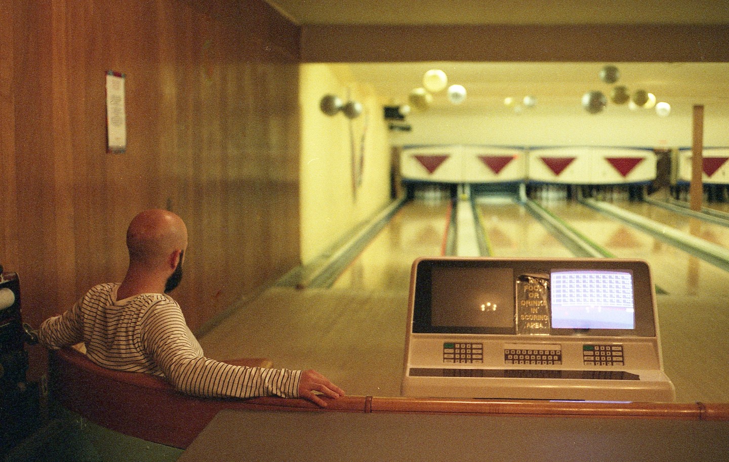 A man with a shaved head sits in a bowling alley, looking towards the lanes.