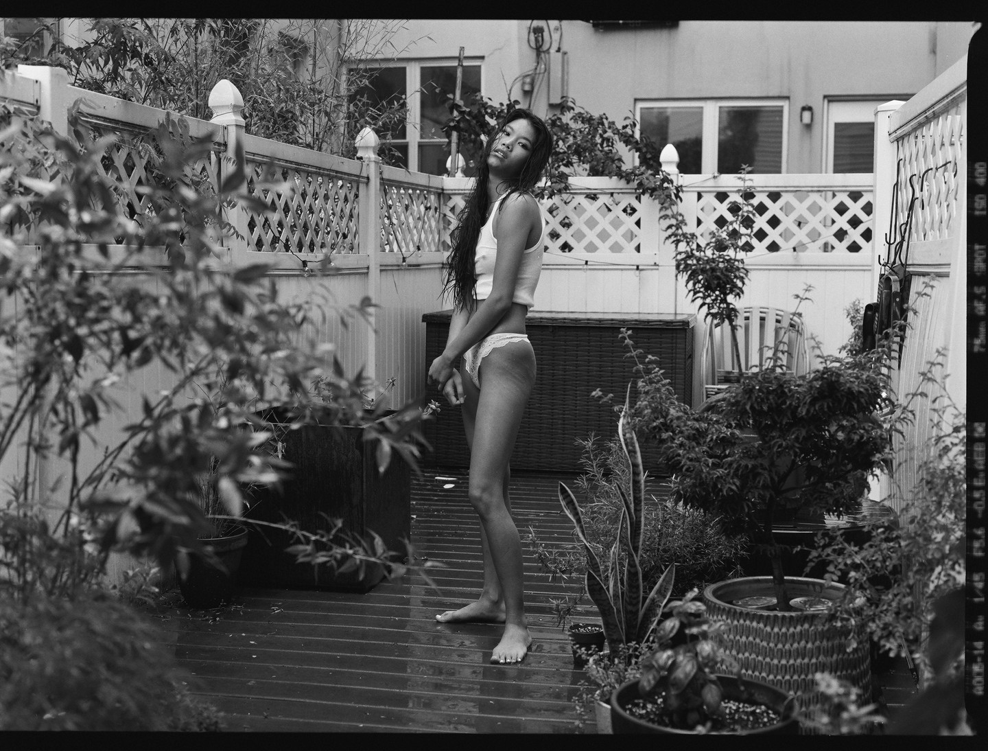 A woman stands barefoot on a wet wooden deck surrounded by potted plants and a white lattice fence.