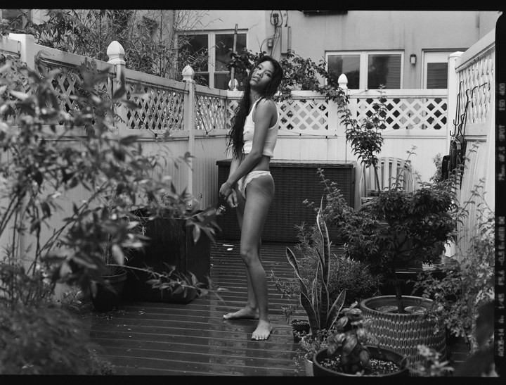 A woman stands barefoot on a wet wooden deck surrounded by potted plants and a white lattice fence.