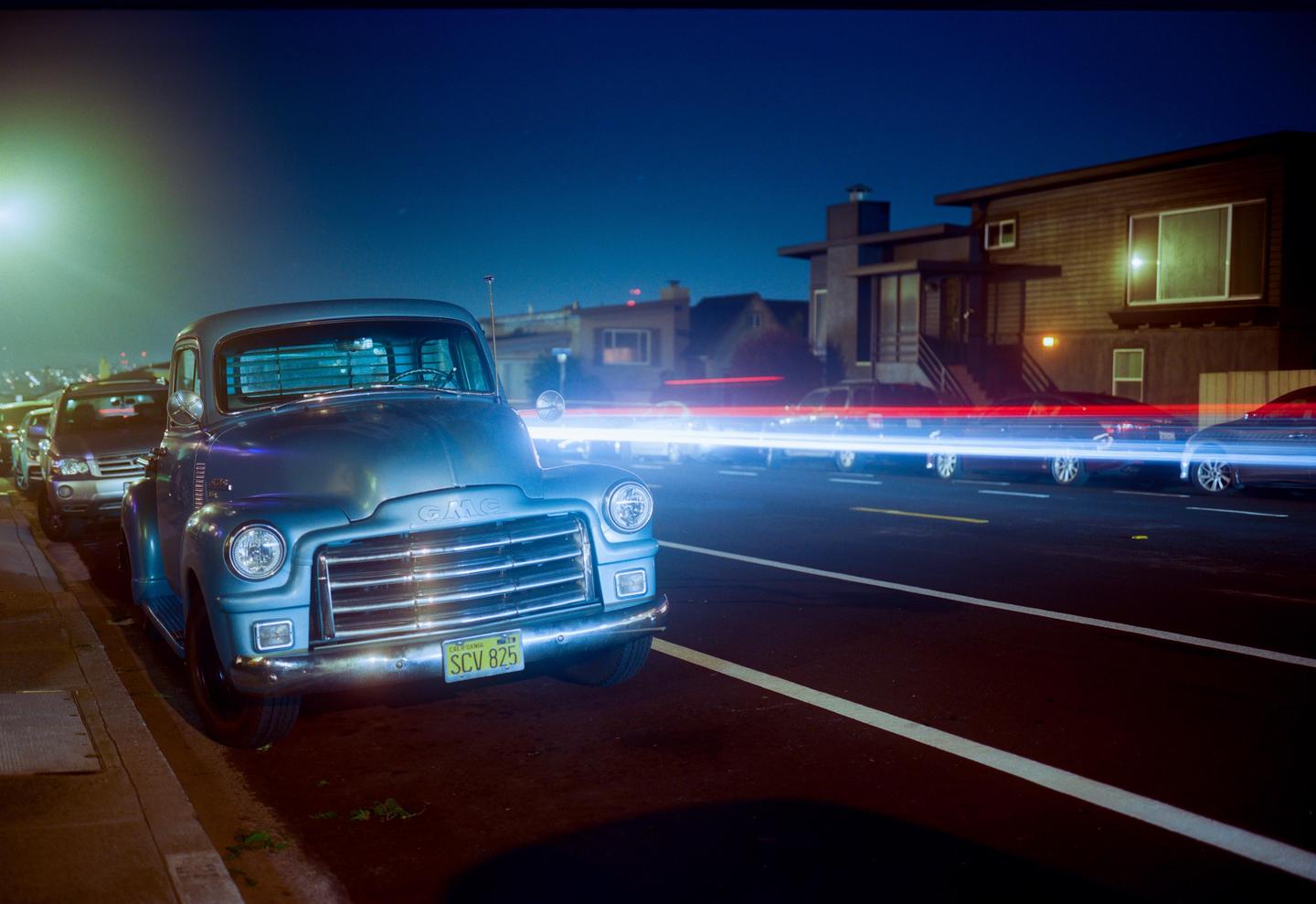 A vintage GMC truck is parked on the side of a street at night with light trails from passing vehicles.