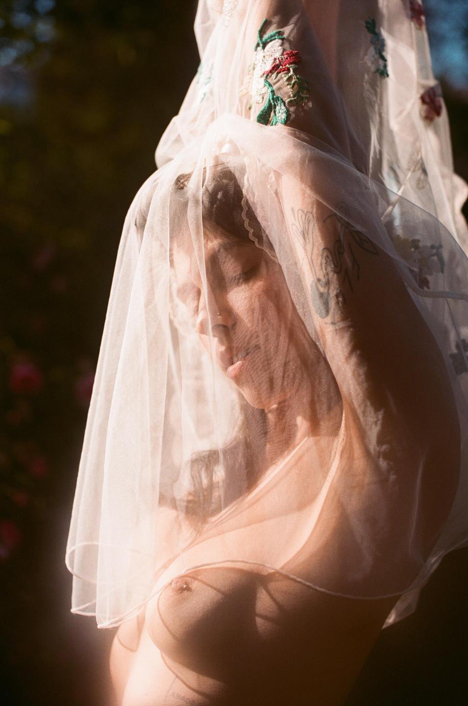 A woman with closed eyes is covered by a sheer veil with embroidered flowers, revealing her bare chest.