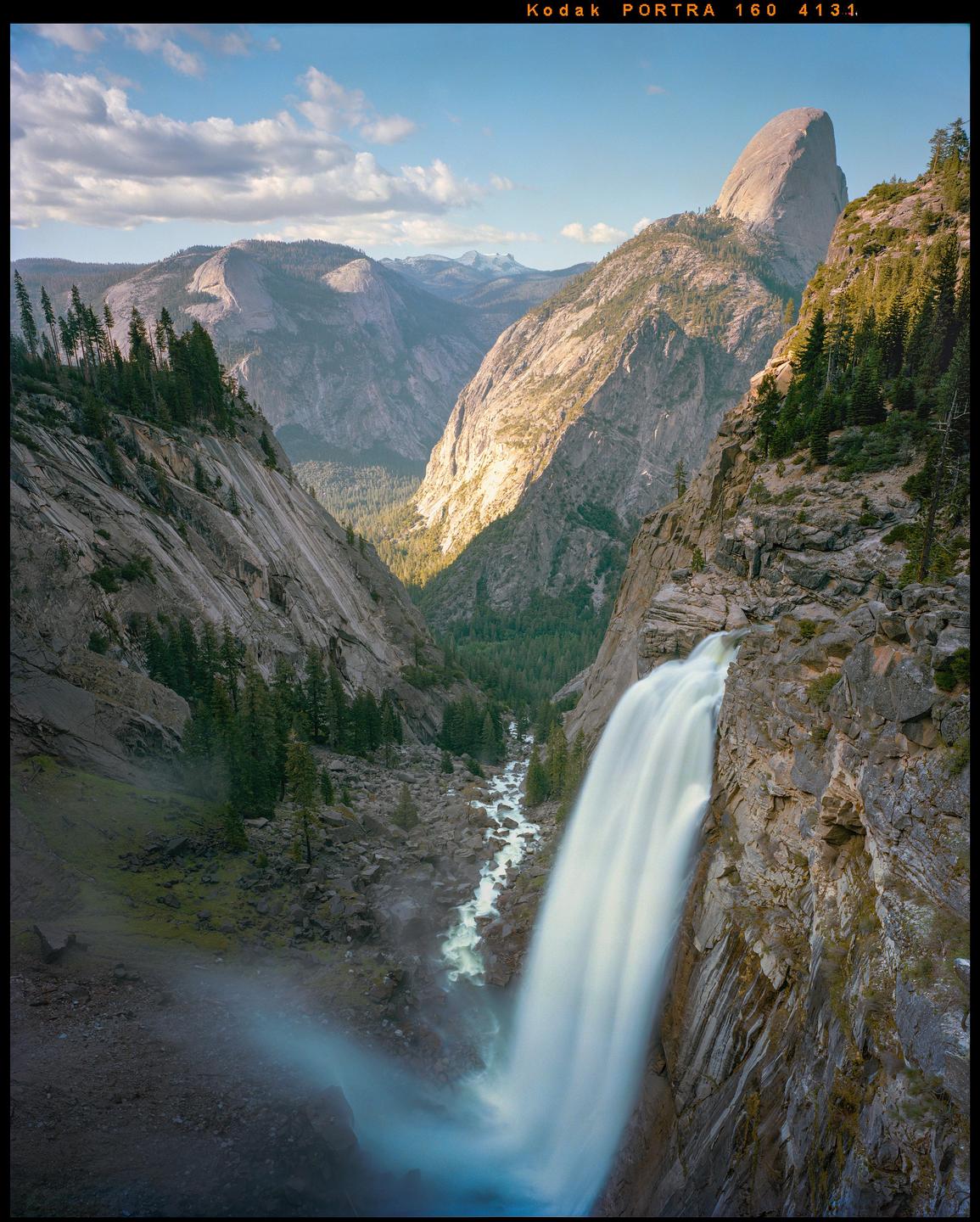 A waterfall cascades down a rocky cliff into a valley with a river and mountains in the background.